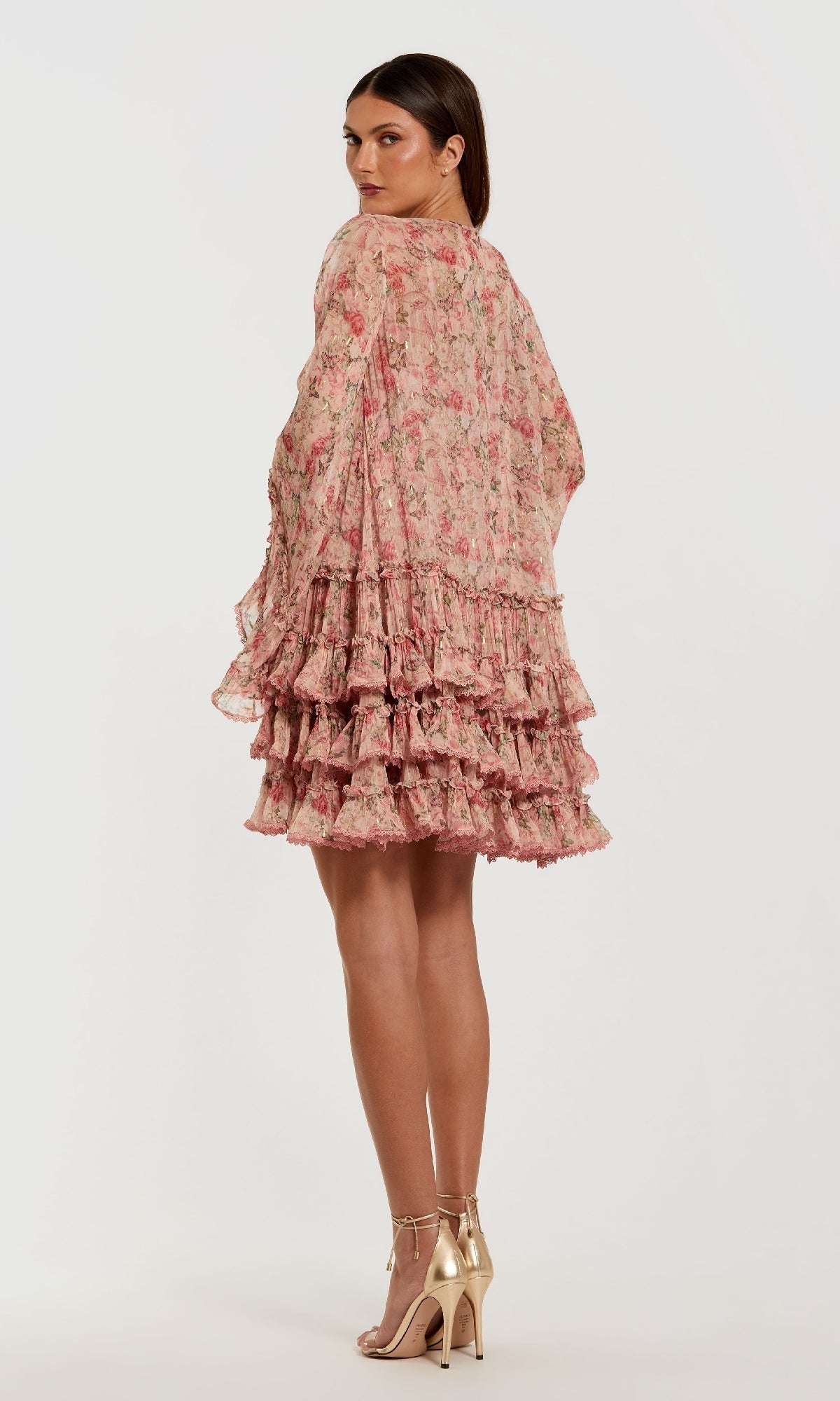 Woman in the Mac Duggal 8336 Print Short Wedding Guest Dress, with long sleeves and a ruffled mini tiered skirt, pairs it with gold high-heeled sandals while standing on a plain background and looking over her shoulder.